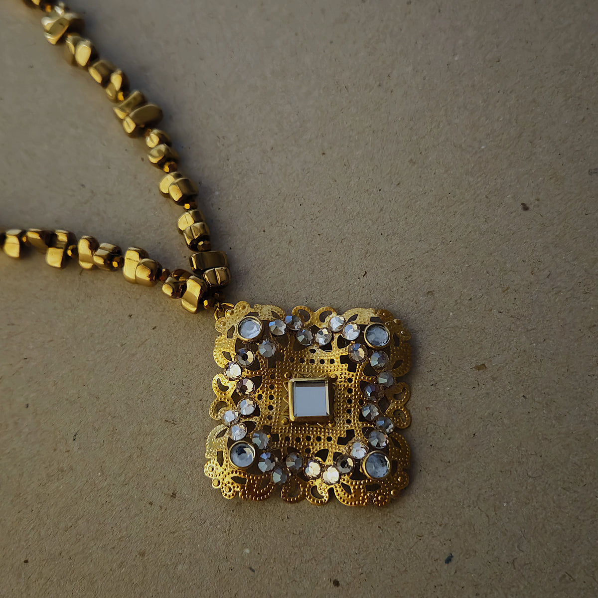 A square gold pendant necklace with intricate filigree detailing, champagne colored rhinestones and kundan style glass